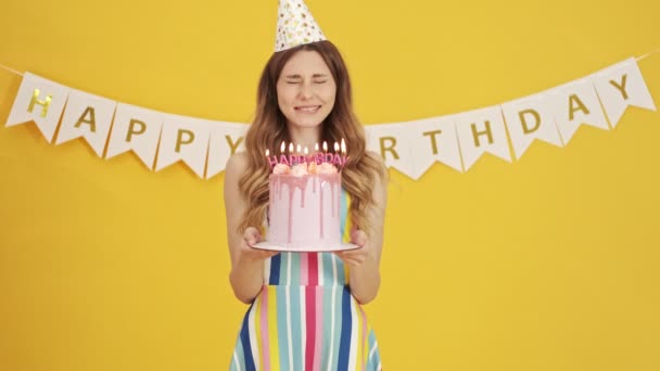 パーティーコーンの幸せな女性は 誕生日ケーキを保持しながら願いを作っていますその後 黄色の背景に隔離されたキャンドルを吹いて — ストック動画