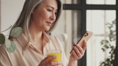 Hoş olgun bir kadın evde akıllı telefonuna bakarken portakal suyu içiyor.