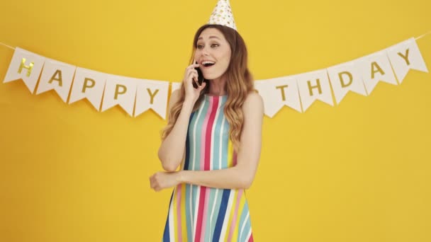 黄色の背景の上に隔離された彼女のスマートフォンの上で話している幸せな誕生日の女性 — ストック動画