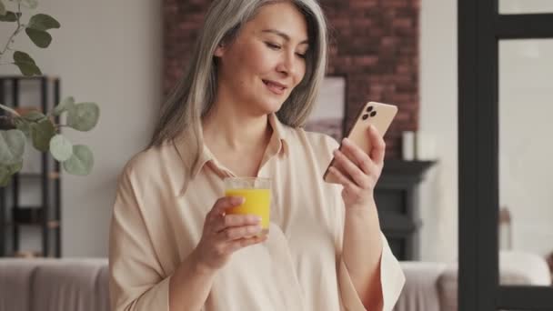 Krásná Zralá Žena Pije Pomerančový Džus Při Použití Svého Smartphonu — Stock video