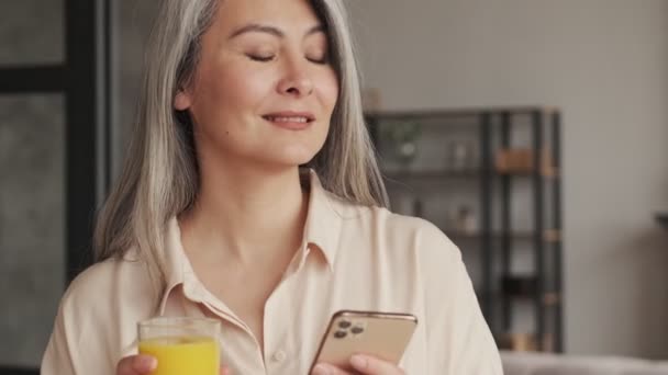 一位心平气和 积极向上的成熟女性在家里厨房里一边看着智能手机里的东西 一边喝着橙汁 — 图库视频影像