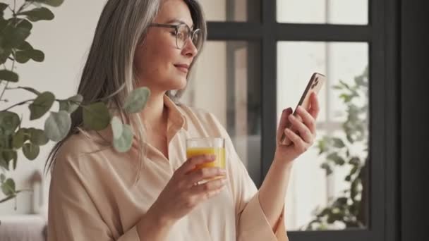 一个戴眼镜的漂亮的成熟女人的侧面形象是 她一边喝着橙汁 一边在家里的厨房里用智能手机看东西 — 图库视频影像