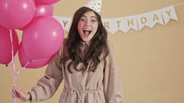 Bir parti konisinde gülümseyen mutlu bir kadın elinde pembe hava balonları ve bej bir arka planda izole edilmiş mutlu bir şekilde bekliyor.