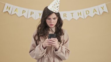 Parti konisindeki üzgün, ağlak bir doğum günü kadını bej arka planda duran akıllı telefonunu kullanıyor.