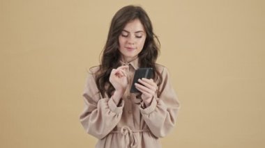 Mutlu bir kadın, bej bir arkaplan üzerinde izole bir şekilde duran akıllı telefonunu kullanırken üzerine bir şey yazmayı seçiyor.