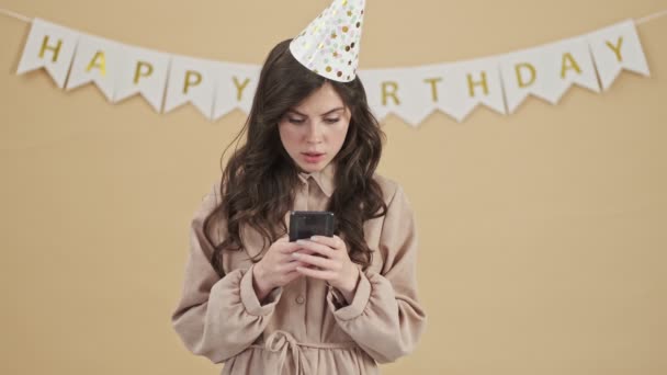 一位专注于生日宴会的女士站在米色的背景上 在智能手机上听到了令人兴奋的好消息 — 图库视频影像