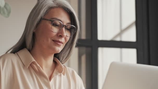 美しいです成熟した女性です眼鏡は作業しています彼女の銀のラップトップコンピュータで家に座って — ストック動画