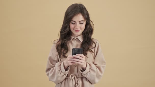一位面带微笑的女士正在用她的智能手机在米色背景下孤立无援地站着 — 图库视频影像