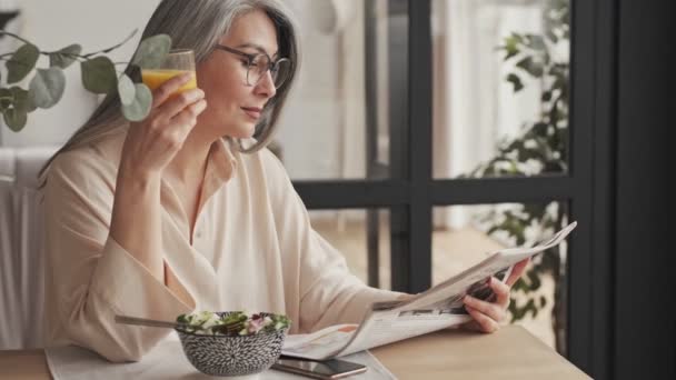 Gorgeous Mature Woman Eyeglasses Reading Newspaper While Eating Breakfast Sitting — стоковое видео