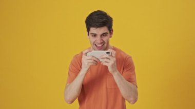 Tişört giyen, akıllı telefondan oynayan ve sarı arka planda sevinen mutlu yakışıklı bir adam.