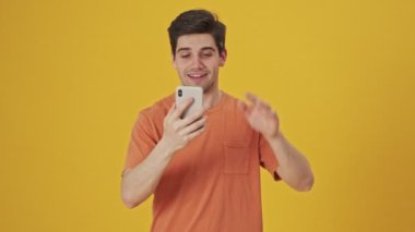 Tişört giyen mutlu yakışıklı adam sarı arka planda akıllı telefondan video konuşmalarıyla konuşuyor.