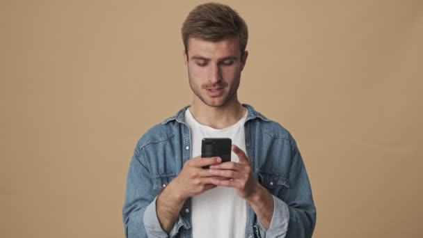 一个专心致志的年轻人站在演播室米色的墙上用智能手机发短信 — 图库视频影像