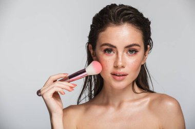 Çekici, seksi, esmer, uzun saçlı bir kadının güzel portresi. Gri arka planda tek başına duruyor. Makyaj fırçası kullanıyor.