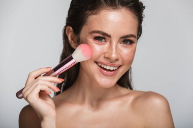 Çekici, gülümseyen, şehvetli, kumral, uzun saçlı bir kadının güzel portresi. Gri arka planda tek başına duruyor, makyaj fırçası kullanıyor.