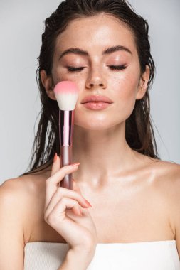 Çekici, seksi, esmer, uzun saçlı bir kadının güzel portresi. Gri arka planda tek başına duruyor. Makyaj fırçası kullanıyor.