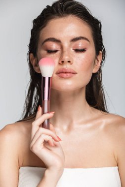 Çekici, seksi, esmer, uzun saçlı bir kadının güzel portresi. Gri arka planda tek başına duruyor. Makyaj fırçası kullanıyor.