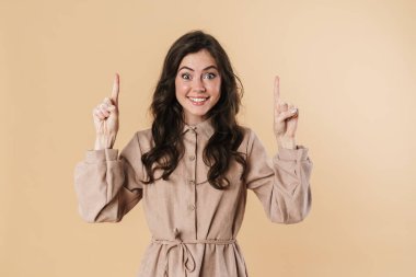 Gülümseyen ve parmaklarını yukarı gösteren neşeli çekici bir kadın görüntüsü bej arka planda izole edilmiş.