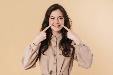 Gülümseyen ve yanaklarını işaret eden neşeli bir kadın bej arka planda izole edilmiş.