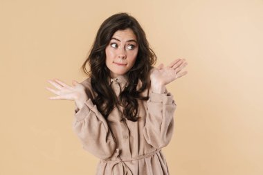 Kafası karışmış, elleri havada poz veren ve bej arka planda izole edilmiş bir kadın resmi.
