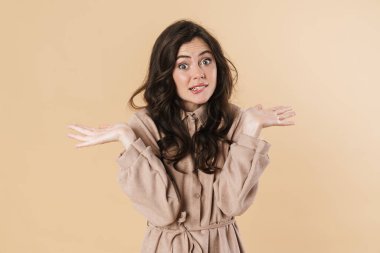 Kafası karışmış, elleri havada poz veren ve bej arka planda izole edilmiş bir kadın resmi.