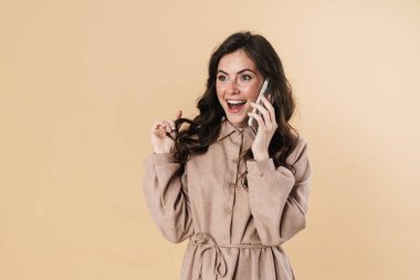 Bej arka planda izole edilmiş, gülümseyen ve cep telefonuyla konuşan neşeli çekici kadın görüntüsü