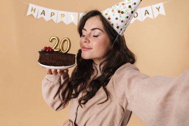 Bej arka planda izole edilmiş doğum günü pastasıyla selfie çeken mutlu bir kadın resmi