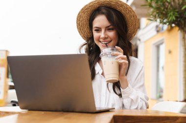 Açık havada rahat bir kafede milkshake içerken dizüstü bilgisayar kullanan gülen genç kadının portresi.