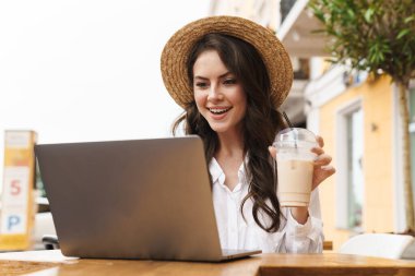 Açık havada rahat bir kafede milkshake içerken dizüstü bilgisayar kullanan gülen genç kadının portresi.