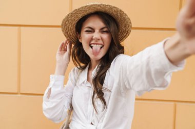 Saman şapkalı, suratını buruşturan ve şehirde yürürken selfie çeken genç bir kadının portresi.