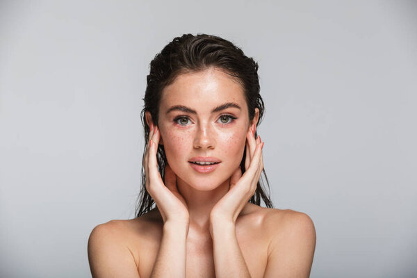 Beauty portrait of an attractive smiling sensual young woman with wet brunette long hair standing isolated over gray background, touching her face, posing