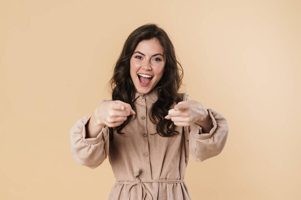 Image of delighted cute woman smiling and pointing fingers at camera isolated over beige background