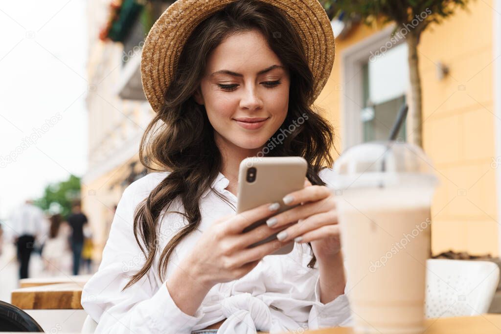 Portrait of young smiling woman using cellphone while drinking milkshake in cozy cafe outdoors