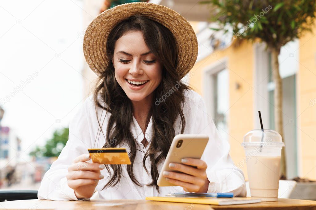 Portrait of young laughing woman using cellphone and holding credit card while sitting in cozy cafe outdoors