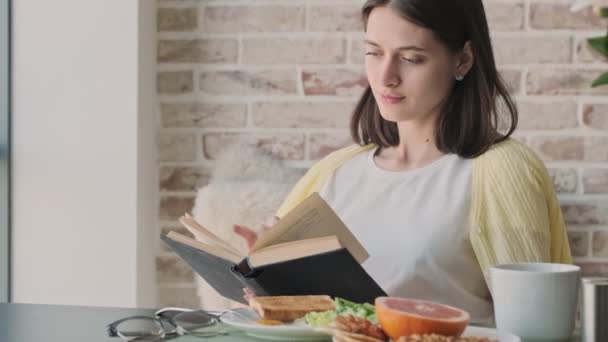 Genç Konsantre Bir Kadın Evde Kitap Okurken Kahvaltı Yapıyor — Stok video