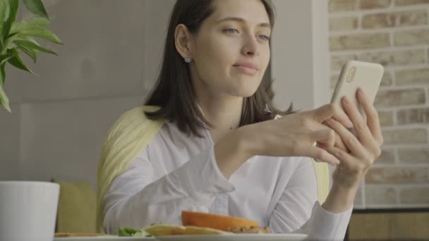 朝食を食べながら携帯電話を使って家の中で喜ぶ若い女性 — ストック動画