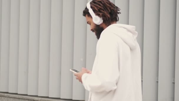 年轻英俊的非洲男子在户外散步 一边用手机 一边用耳机听音乐 — 图库视频影像