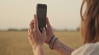 Elbise giyen, buğday tarlasında akıllı telefondan fotoğraf çeken çekici kadının yakın görüntüsü.