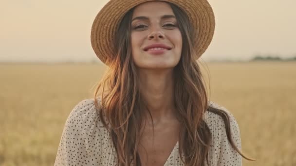 遊び心のある魅力的な女性は ドレスやスタイリッシュな帽子をかぶって彼女の髪で遊んで 小麦畑にポーズをとりながらカメラを見て — ストック動画