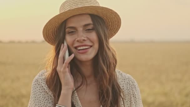 小麦畑を歩いている間にスマートフォンで話すドレスとスタイリッシュな帽子を身に着けている陽気な魅力的な女性 — ストック動画