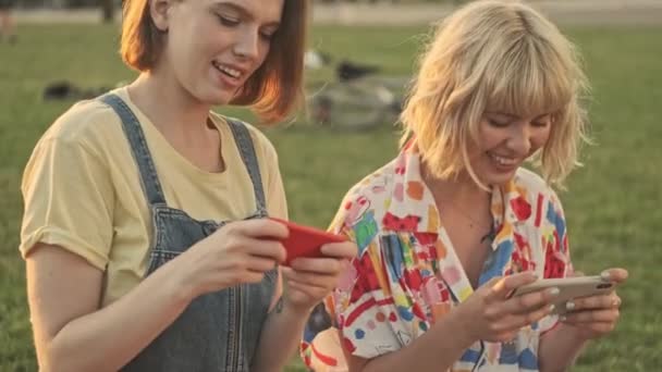2人の美しい笑顔の若い女の子が公園の芝生の上に座っているスマートフォンでデジタルゲームをしています — ストック動画