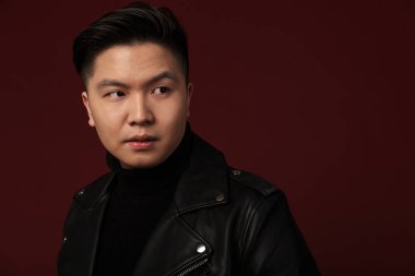 Deri ceketli ciddi bir Asyalı adam resmi. Burgonya arka planında izole edilmiş bir şekilde poz veriyor ve bakıyor.