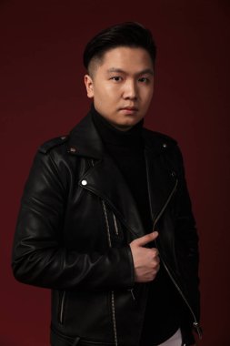 Deri ceketli ciddi bir Asyalı adamın fotoğrafı. Burgonya arka planında izole edilmiş kameraya bakıyor.