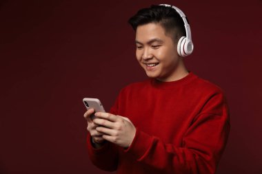 Burgonya arka planında izole edilmiş kulaklık ve cep telefonu kullanan neşeli Asyalı genç adam görüntüsü
