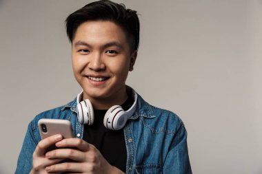 Gri arka planda izole edilmiş akıllı telefon ve kablosuz kulaklık kullanan gülümseyen Asyalı genç adam görüntüsü