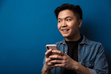Mavi arka planda izole edilmiş, cep telefonu kullanan günlük kıyafetler giyen neşeli Asyalı bir adamın portresi.