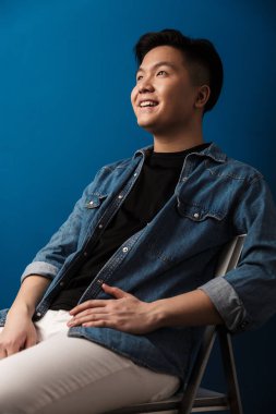 Mavi arka planda izole edilmiş bir sandalyede otururken gülümseyen heyecanlı Asyalı adamın görüntüsü.