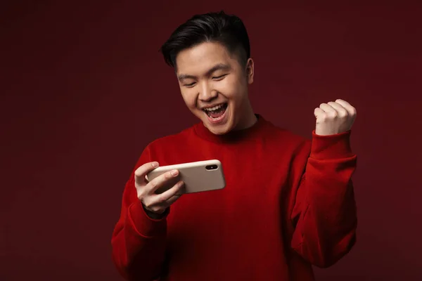 バーガンディを背景に隔離された携帯電話を使用している間 興奮したアジア人男性が勝者のジェスチャーを作るイメージ — ストック写真