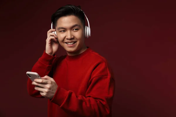 バーガンディを背景に孤立したヘッドフォンや携帯電話を使用している間に笑顔楽しいアジアの若者のイメージ — ストック写真