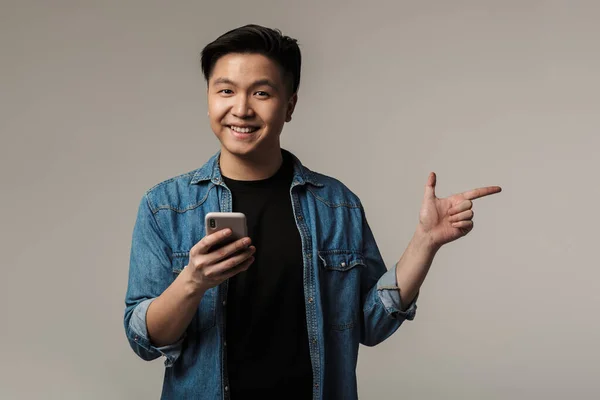Image of smiling asian man pointing finger aside and using smartphone isolated over gray background