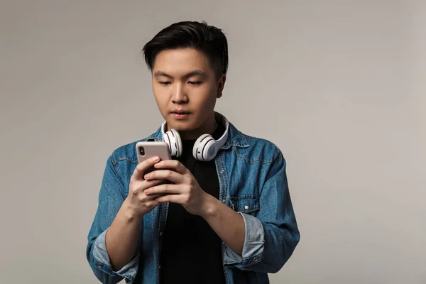 灰色の背景に隔離されたスマートフォンやワイヤレスヘッドフォンを使用して焦点を当てた若いアジア人の男のイメージ — ストック写真